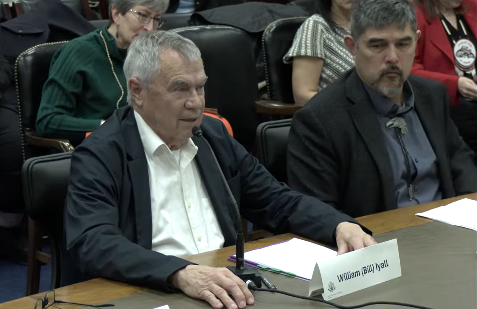 A seated speaker addresses a microphone at a long desk during a formal meeting in Washington, D.C. Papers and a name placard reading “William (Bill) Iyall” rest on the table, with several attendees seated behind and beside the speaker.