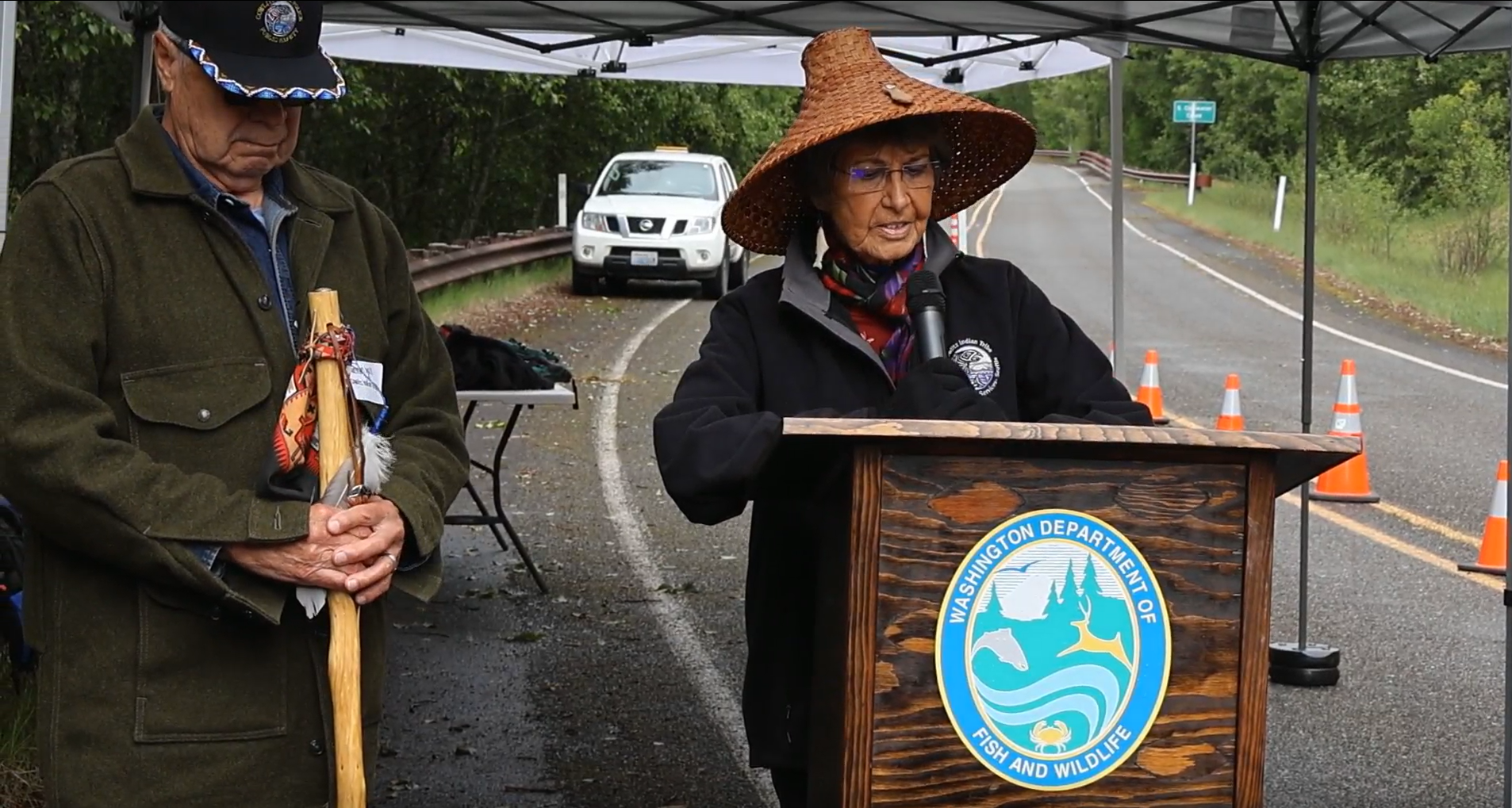 Cowlitz Indian Tribe Spiritual Leader speaking in front of podium labeled Washington Department of Fish and Wildlife with Cowlitz Chairman on her right looking down with a cowlitz indian tribe logo on his hat and a traditional walking stick in his arm. Both appearing under a pop-uptent. 