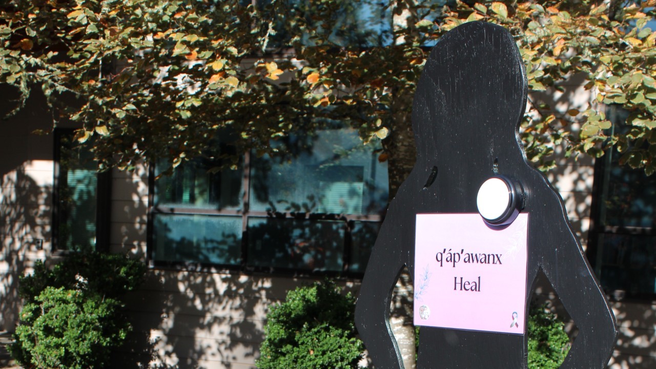 A silhouette display stands outdoors with a sign that reads in Cowlitz Language “q̓áp̓awnx – Heal.” A pot of red flowers is placed at the base of the display as part of a domestic violence awareness exhibit.
