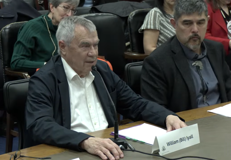 A seated speaker addresses a microphone at a long desk during a formal meeting in Washington, D.C. Papers and a name placard reading “William (Bill) Iyall” rest on the table, with several attendees seated behind and beside the speaker.