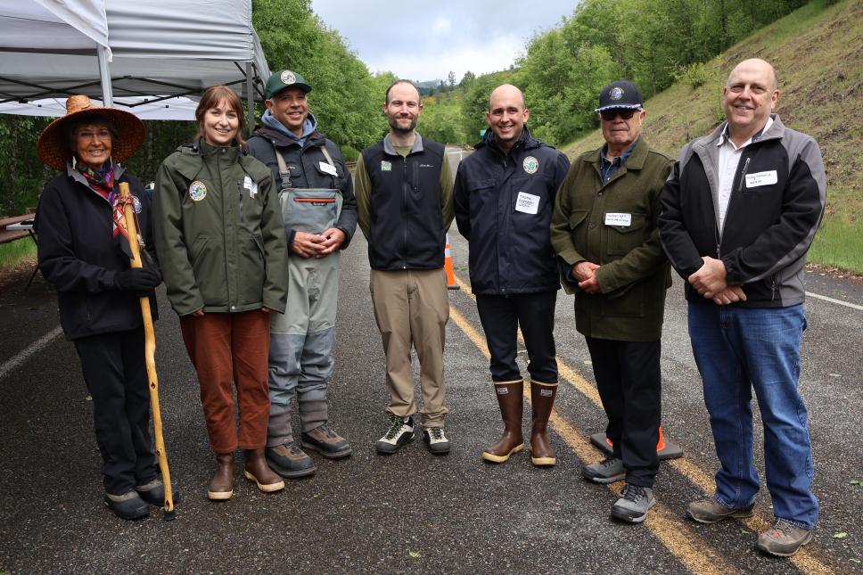 Seven officials from Cowlitz Indian, WDFW in outdoor field gear pose for a photo on a paved road beside a canopy tent, with a forested hillside in the background.
